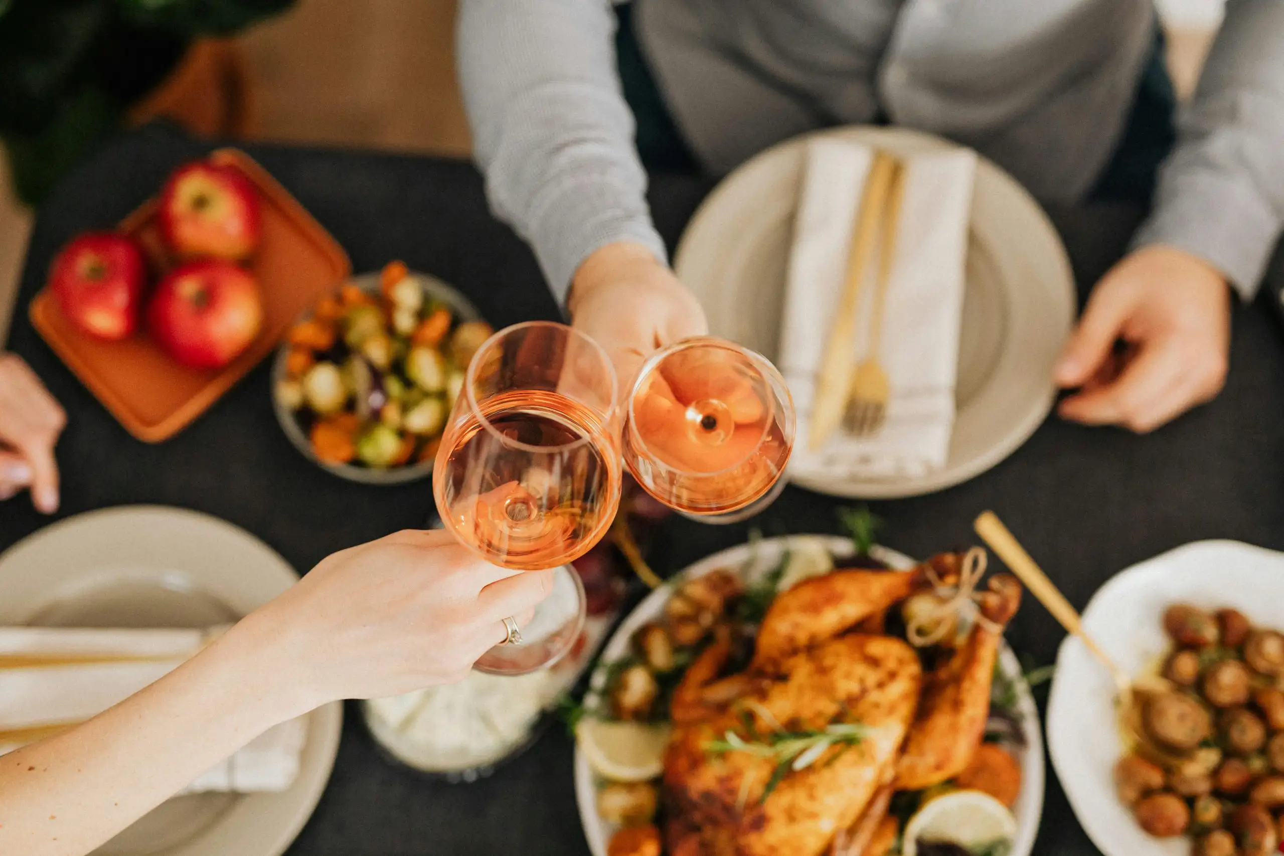 Two people clinking glasses of rosé wine over a holiday dinner table with roasted chicken and side dishes.