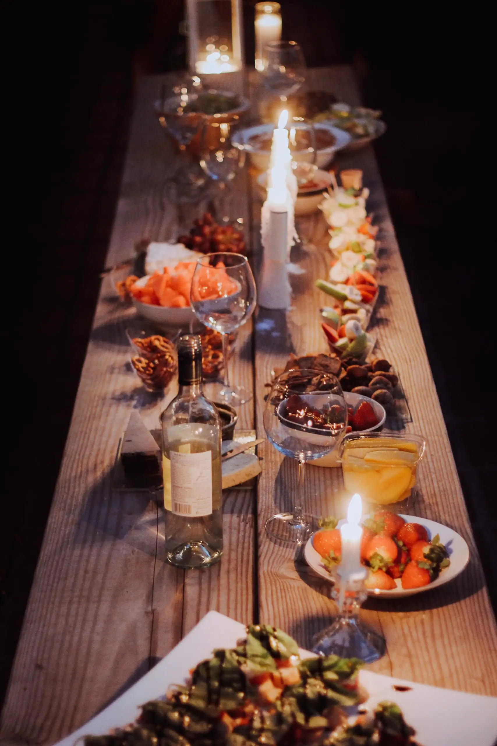 Long wooden dinner table with candles, wine, cheese, fruit, and appetizers.