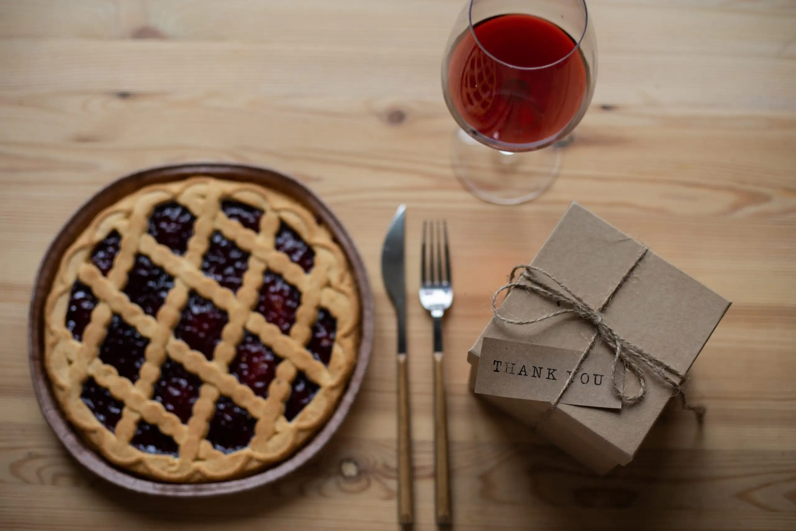 Don't Miss These Wine and Dessert Pairings | Domenico Winery A wooden table setting featuring a lattice-top cherry pie, a glass of red wine, and a brown kraft paper gift box with a "Thank You" tag.