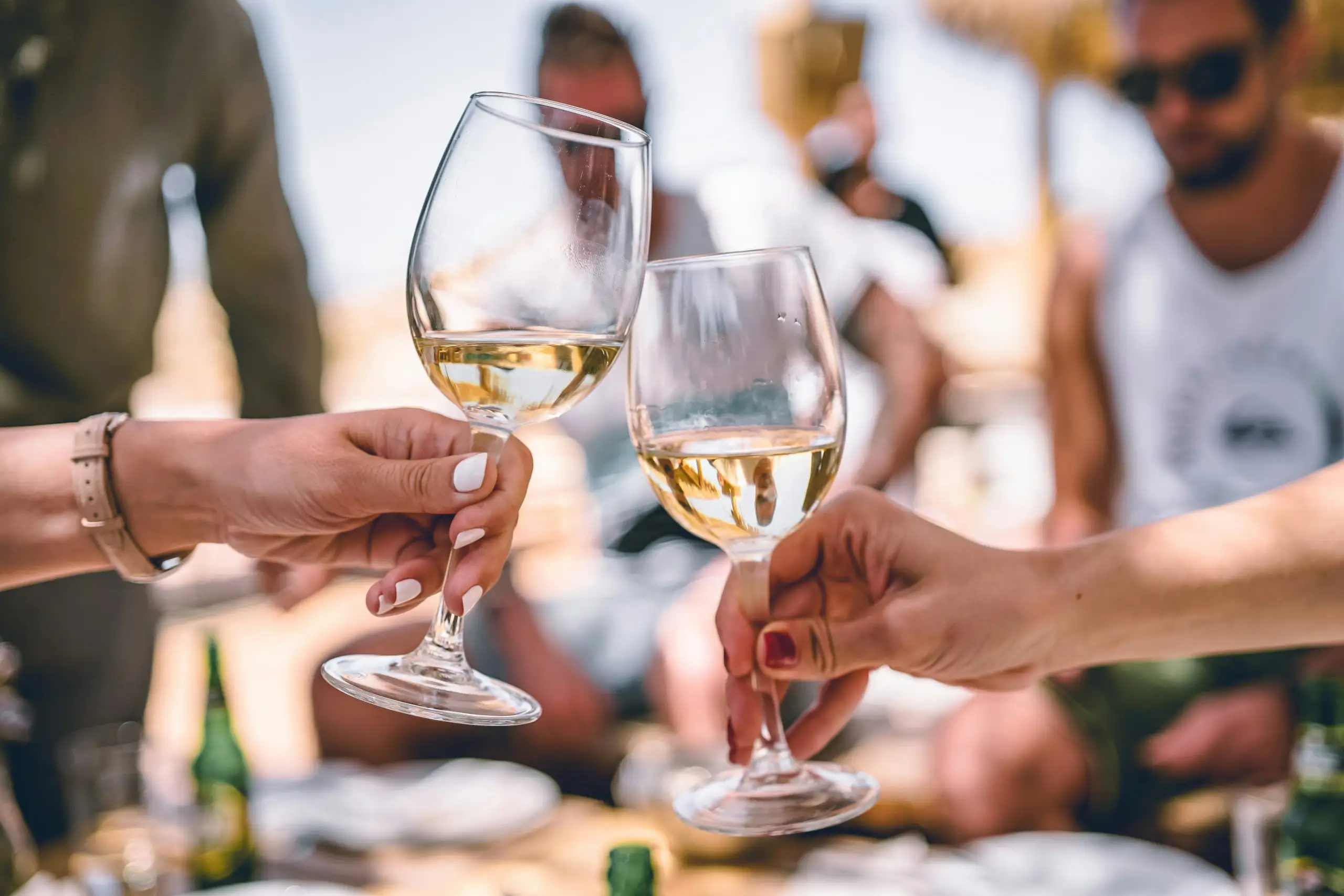 Two hands clinking glasses of white wine in a celebratory toast at a sunny outdoor gathering.