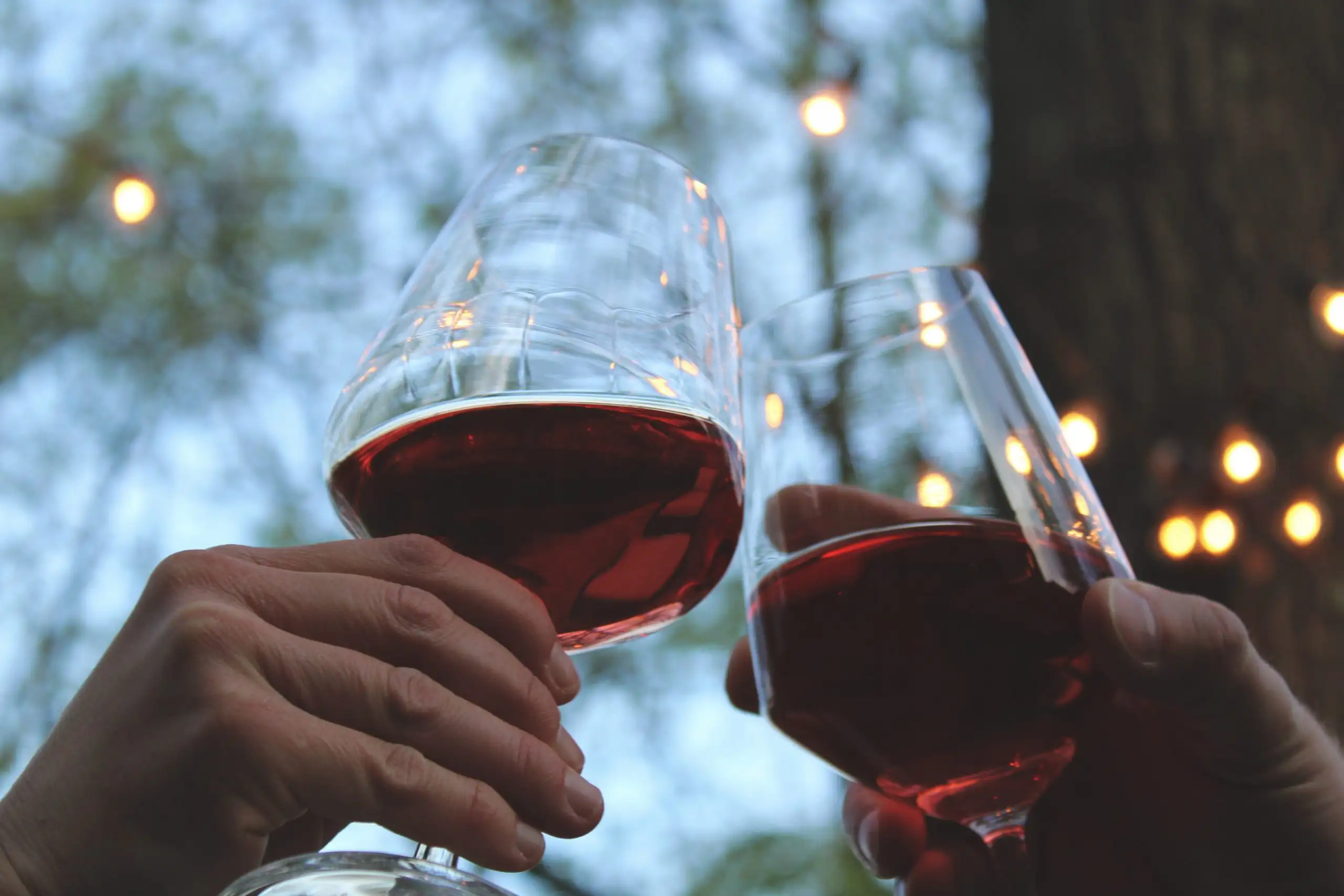 Two people clinking glasses of red wine outdoors in the evening with soft string lights in the background.