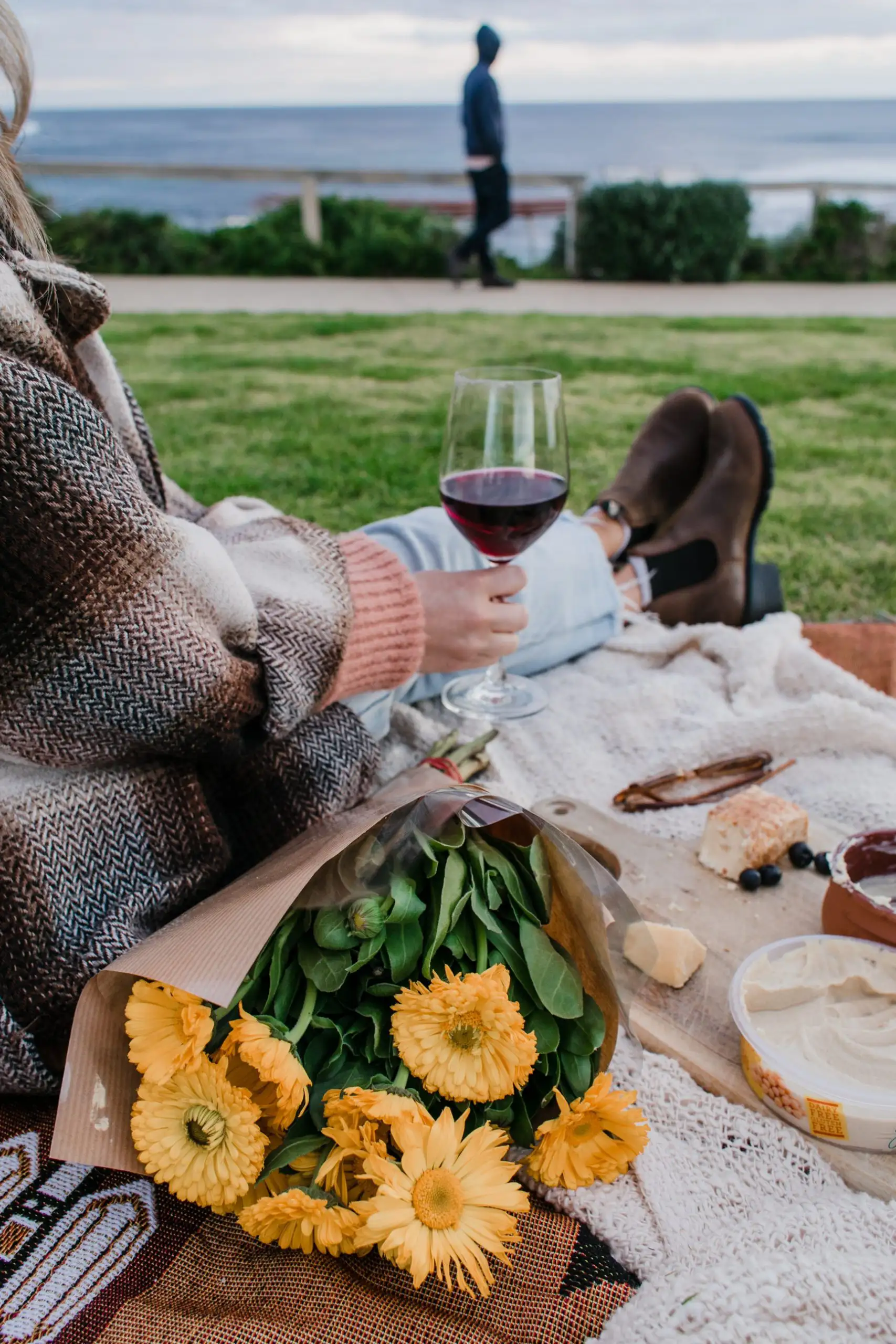 Person relaxing on a blanket near the ocean holding a glass of red wine, with yellow flowers and picnic foods in the foreground.