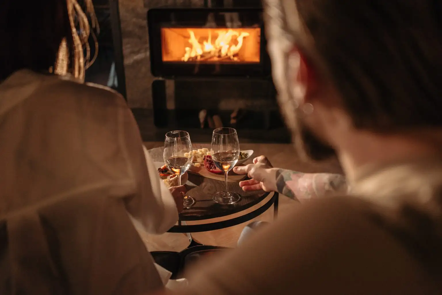 A couple viewed from behind sitting in front of a glowing wood-burning stove with wine glasses and a snack board on a small table.