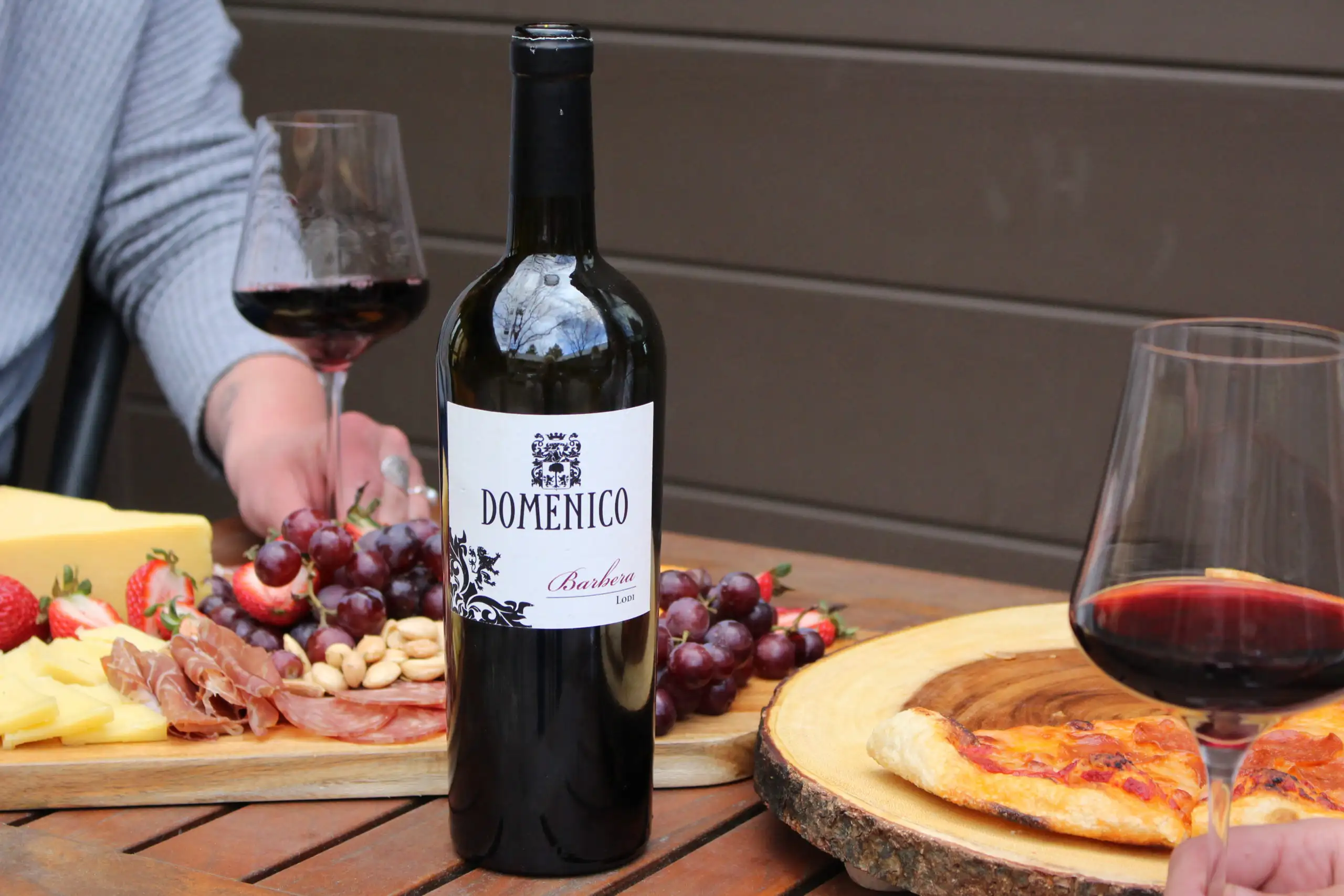 5 Springtime Wines You Must Try | Domenico Winery Bottle of Domenico Barbera red wine on a wooden table with glasses of red wine, a charcuterie board with cheese, grapes, and cured meats, and a slice of pizza.