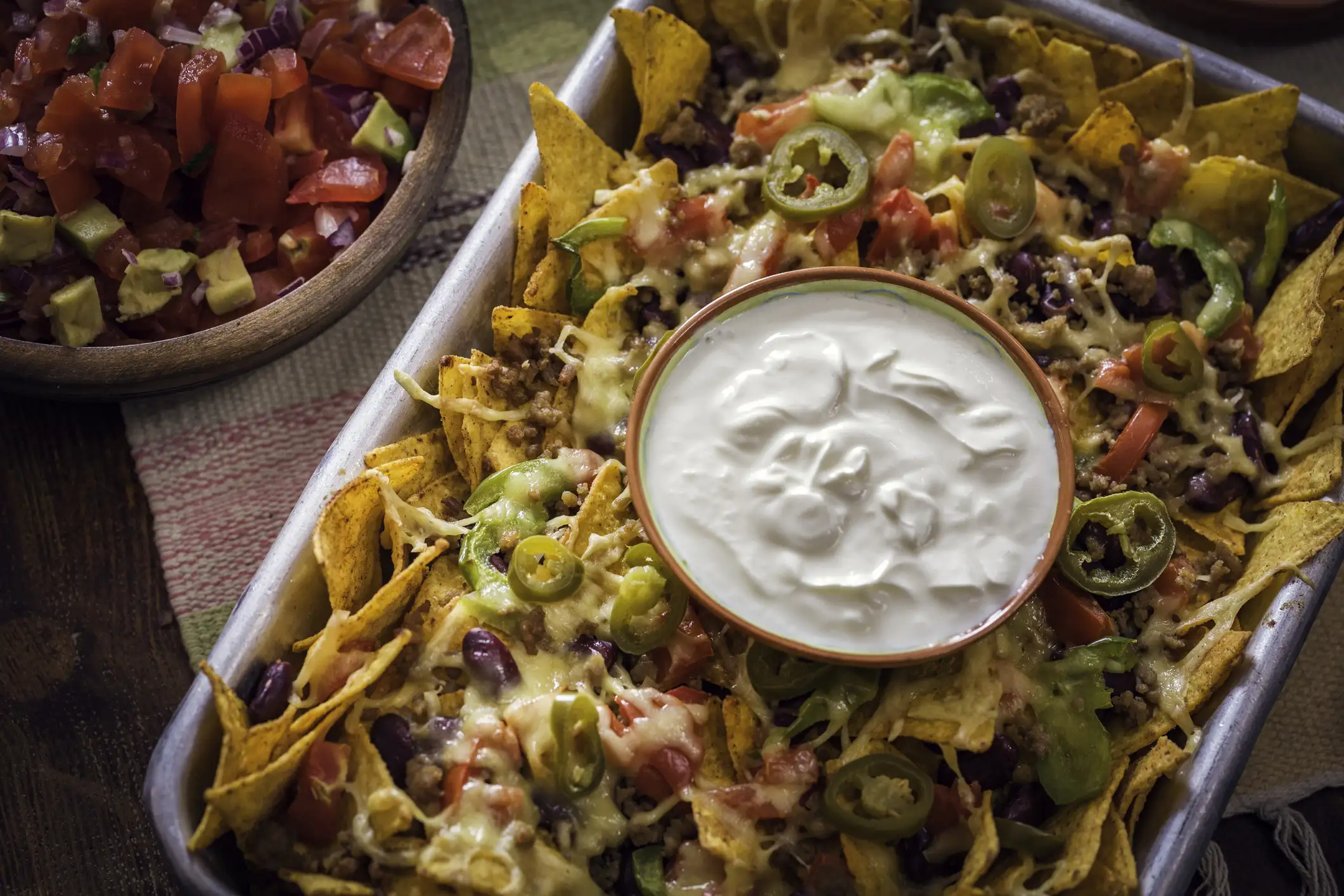The Ultimate Super Bowl Wine Pairings | Domenico Winery A metal tray filled with corn tortilla chips topped with melted cheese, ground beef, black beans, jalapeños, and peppers, served with a side bowl of fresh tomato and avocado salsa.
