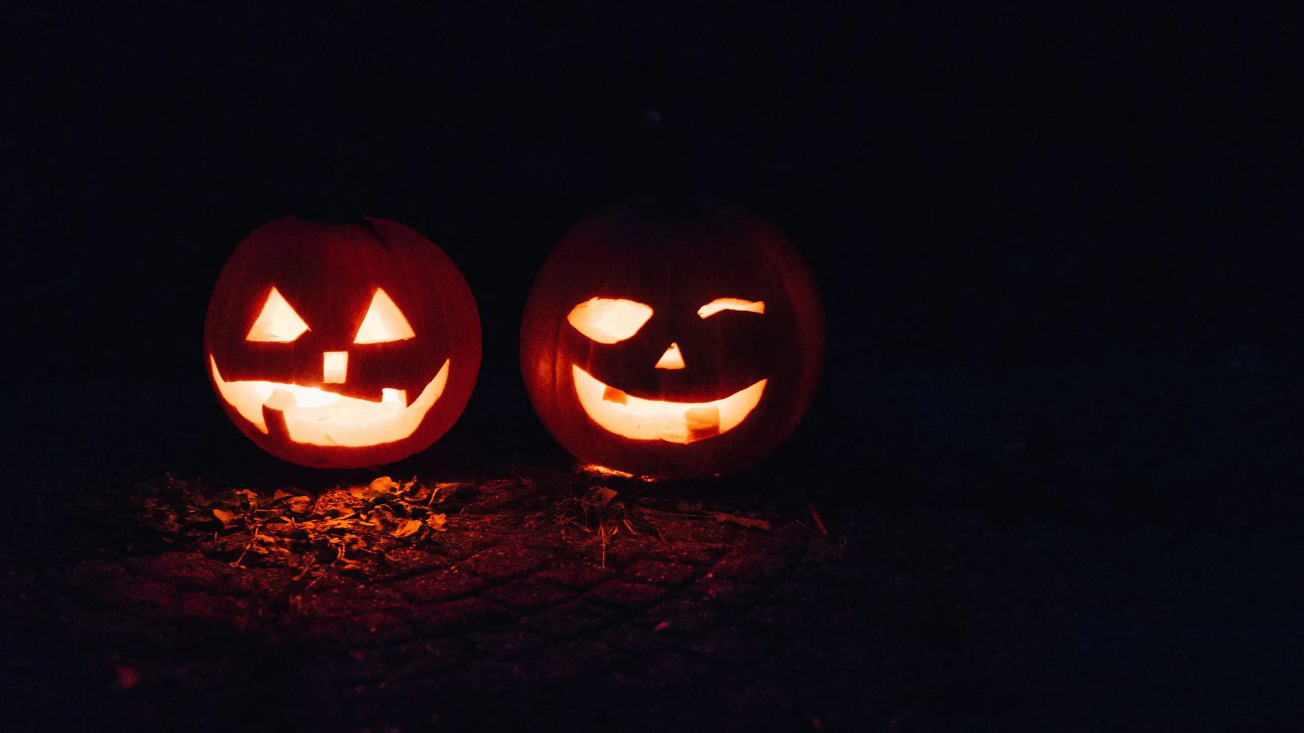 Two carved Halloween pumpkins, one with a smiling face and the other with a winking face, lit up against a dark background.