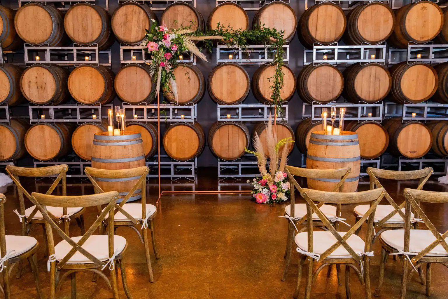 An indoor event space set up for a ceremony with wooden cross-back chairs, a copper pipe arch decorated with pink flowers, and a background of stacked wine barrels.