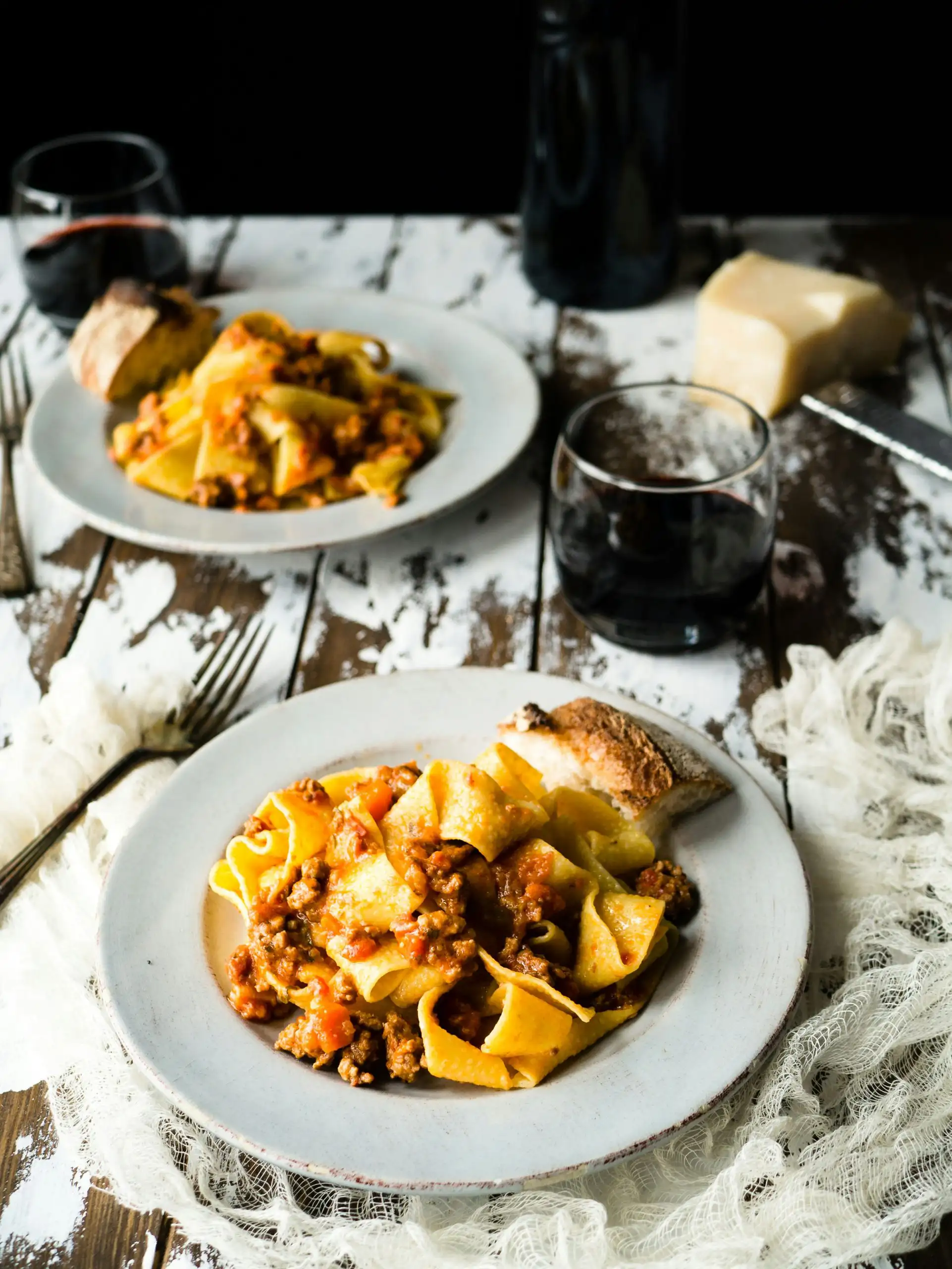 Plate of pappardelle pasta with bolognese sauce on a rustic wooden table with red wine, bread, and cheese.