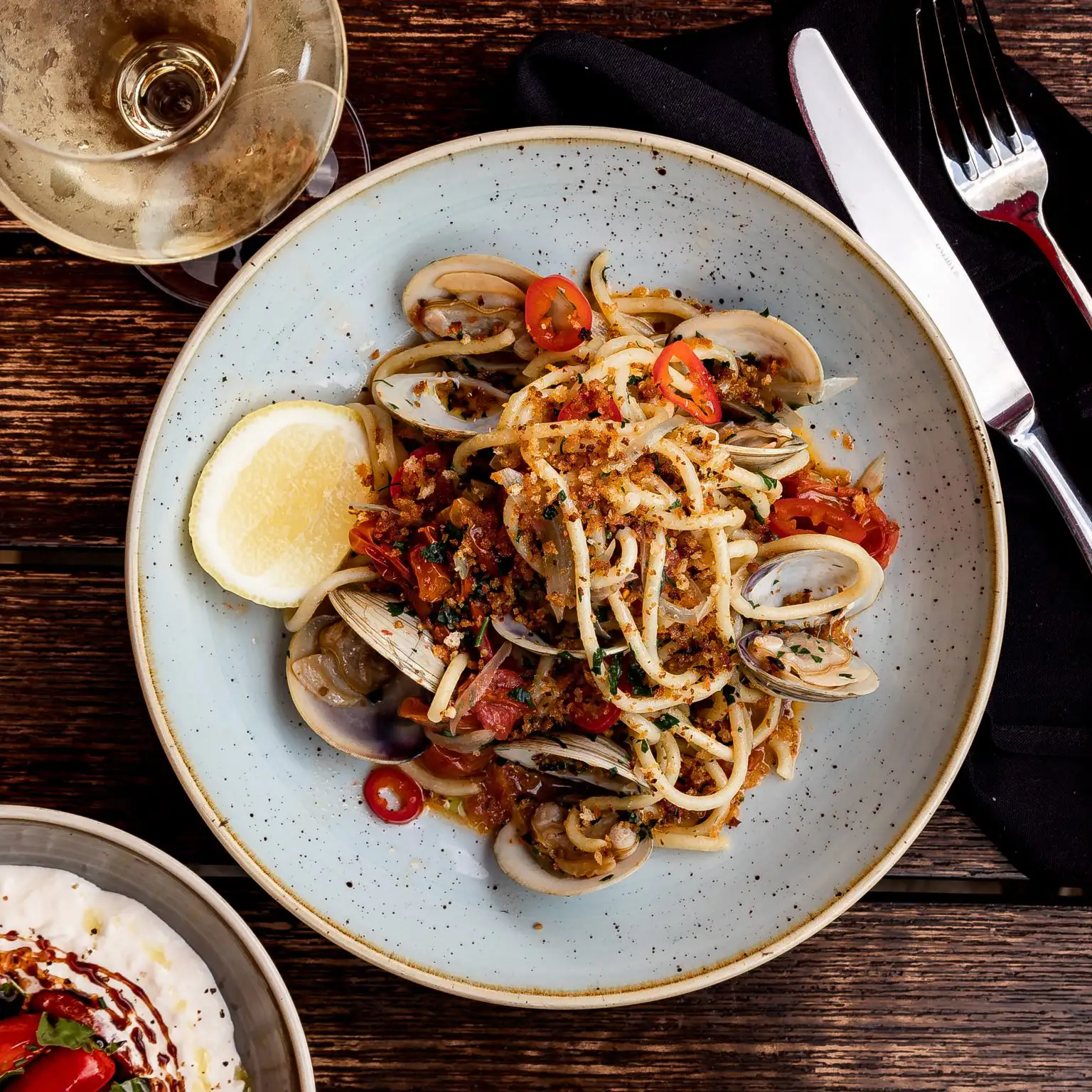 Italian Christmas Traditions Everyone Should Know | Domenico Winery A top-down view of a bowl of linguine pasta with clams, sliced red chilies, breadcrumbs, and a lemon wedge on a dark wood table.