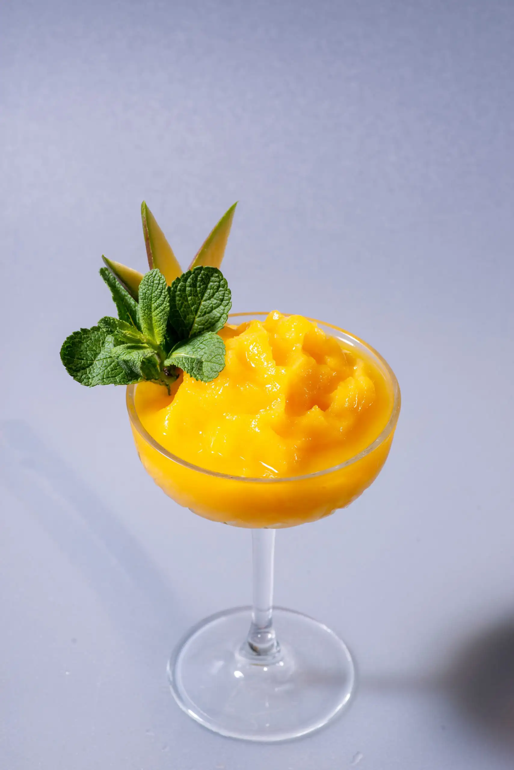 Don't Miss These Wine and Dessert Pairings | Domenico Winery Vibrant yellow mango sorbet in a crystal coupe glass, garnished with fresh mint leaves and a thin fruit slice against a grey background.