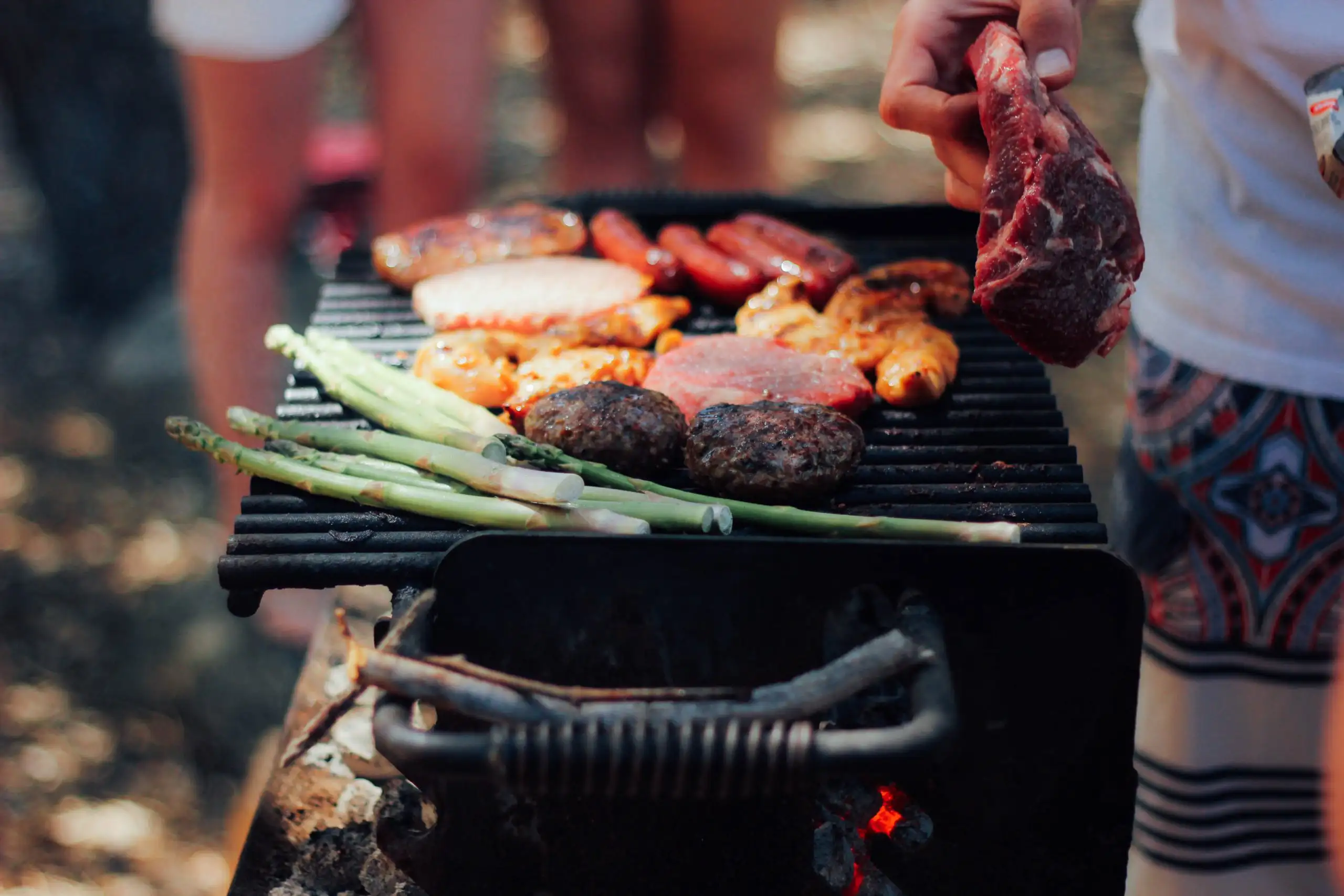 Person placing raw steak on a grill with burgers, sausages, chicken, and asparagus cooking outdoors.