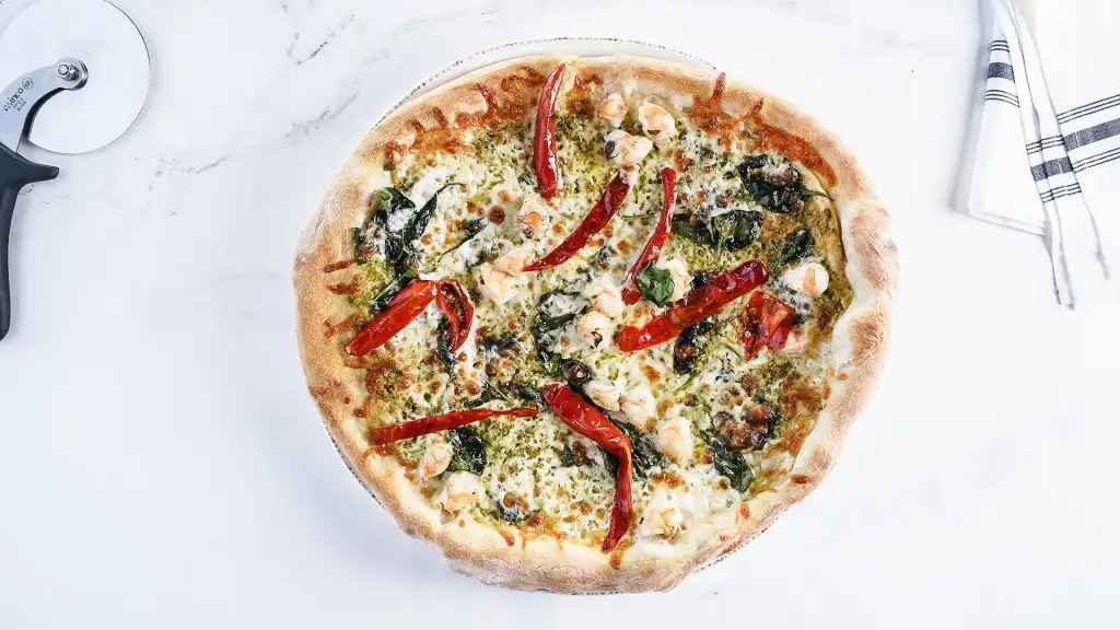 How to Pair Wine with Pizza (Guide) | Domenico Winery Pesto pizza topped with sun-dried tomatoes, spinach, and shrimp, served with a pizza cutter on a marble countertop.