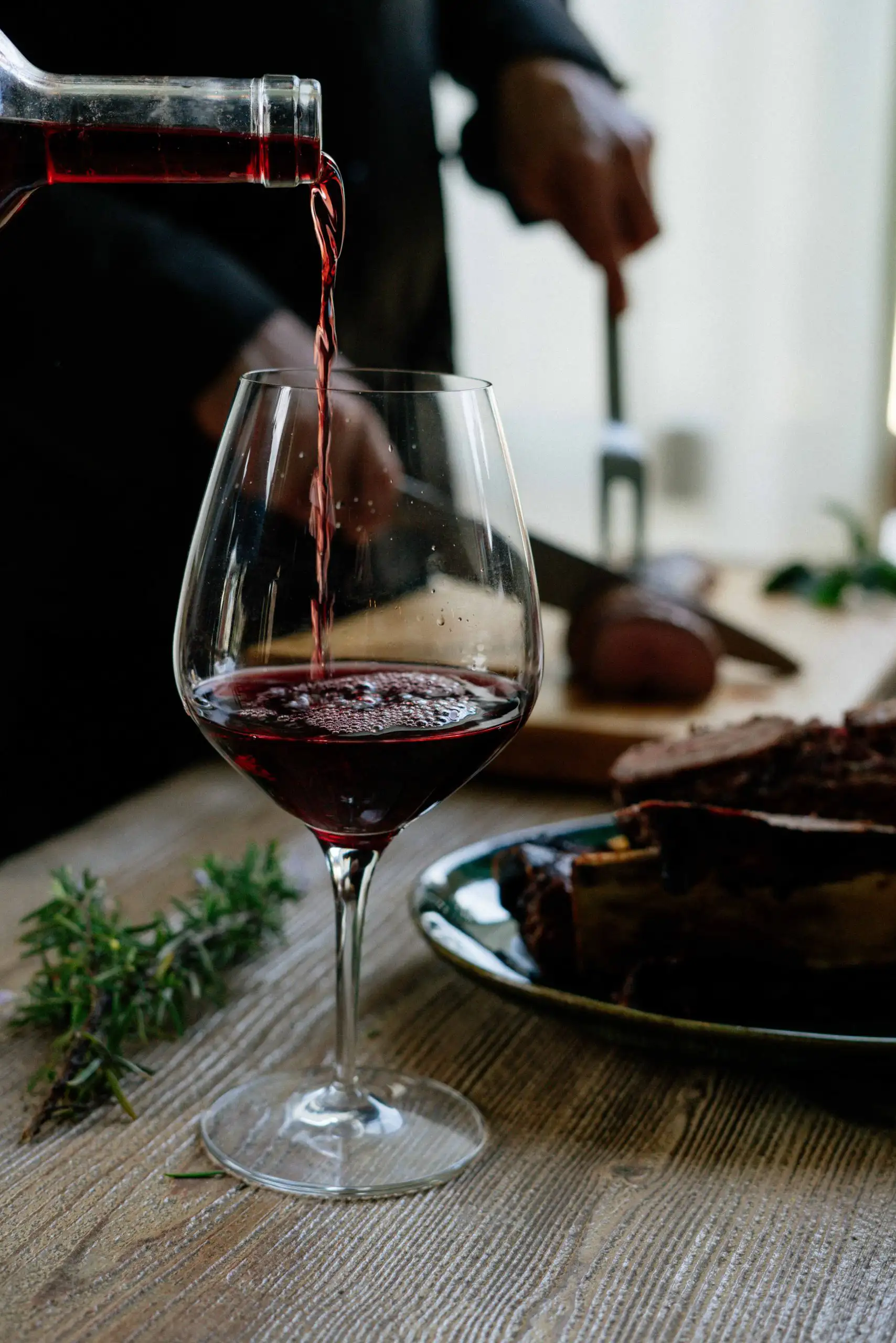 A glass of red wine being poured with a steak and rosemary on a wooden table.