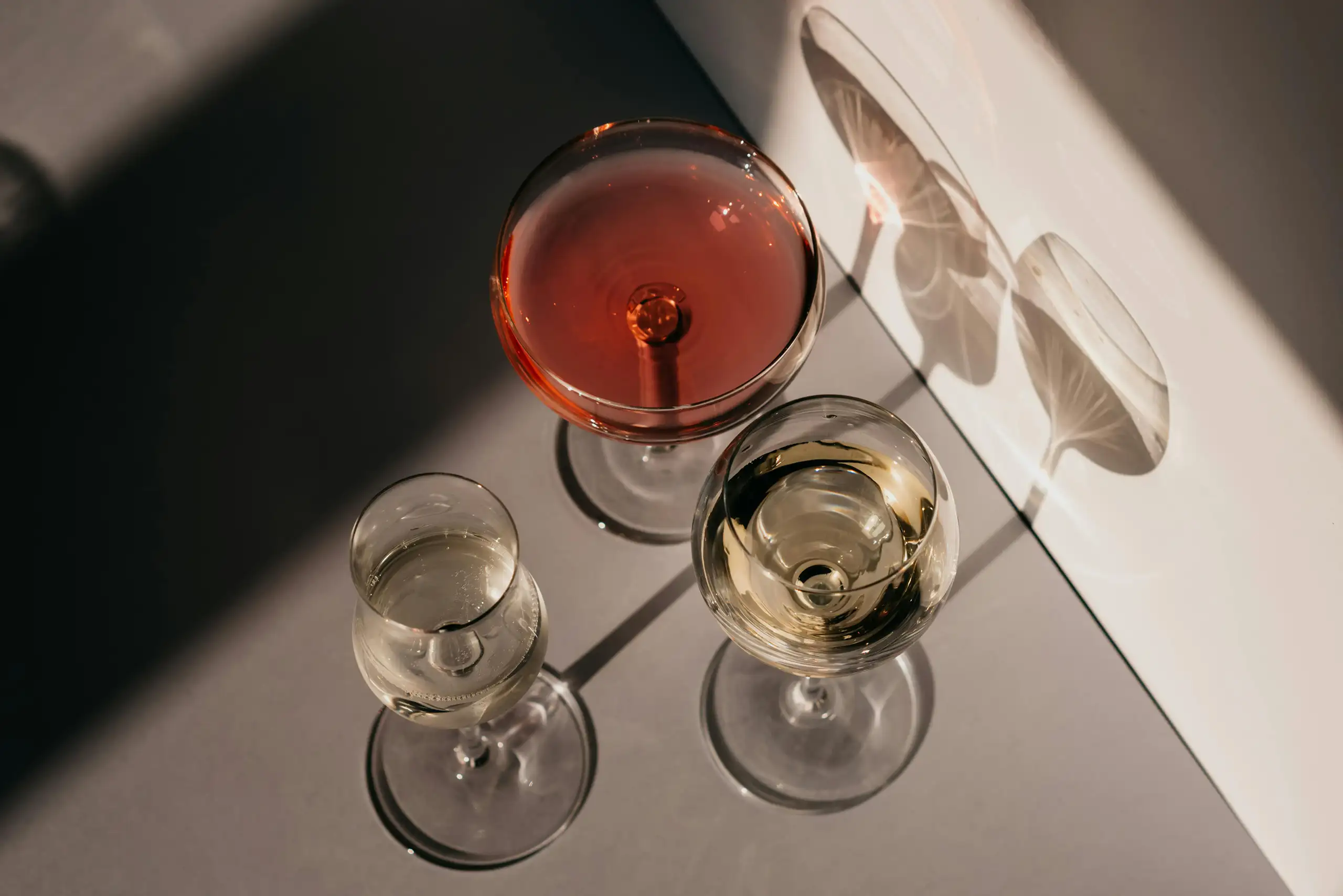 Three wine glasses with rosé and white wine on a table, illuminated by sunlight casting shadows on a wall