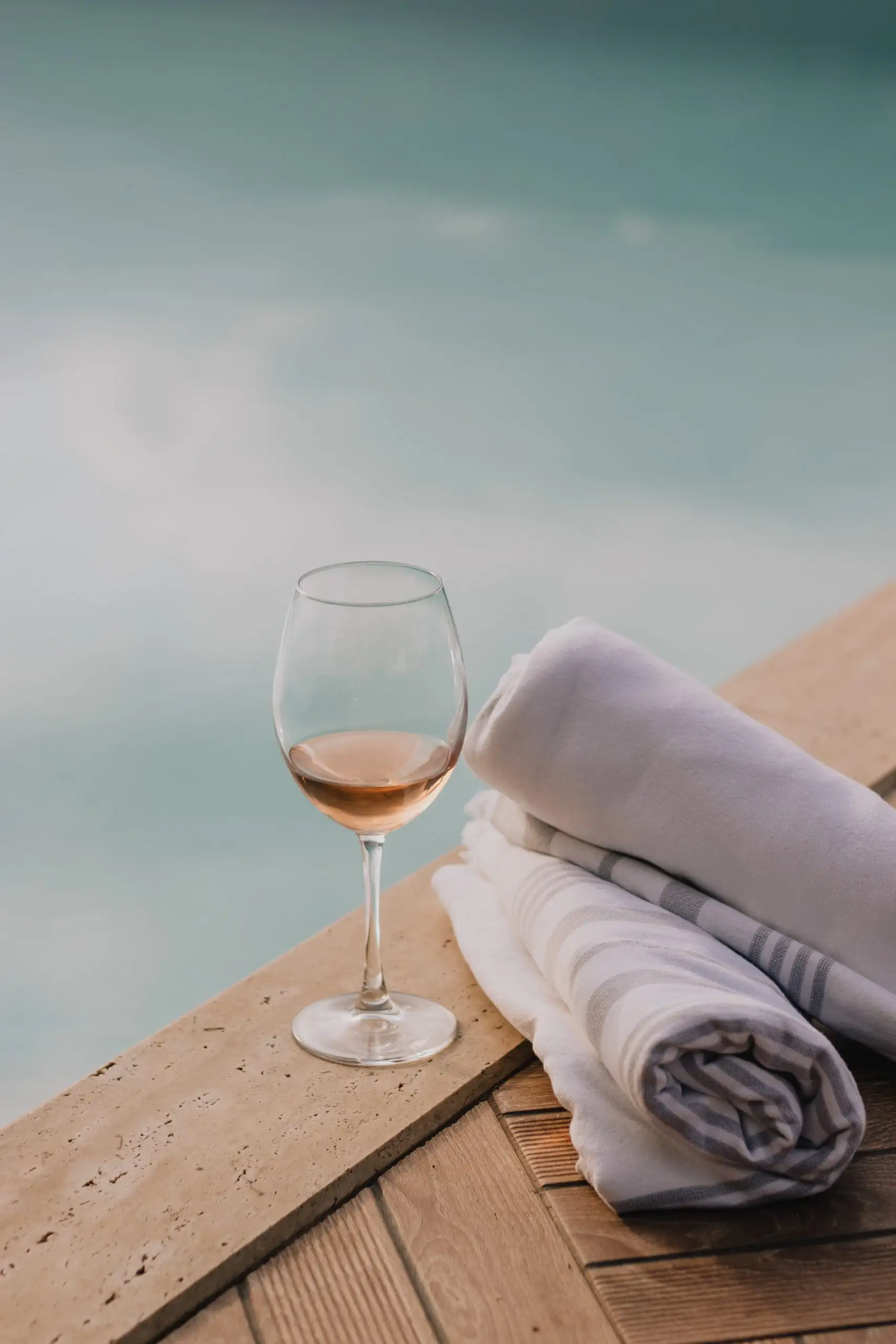 A glass of rosé wine placed on a stone surface next to rolled-up white towels by a pool.