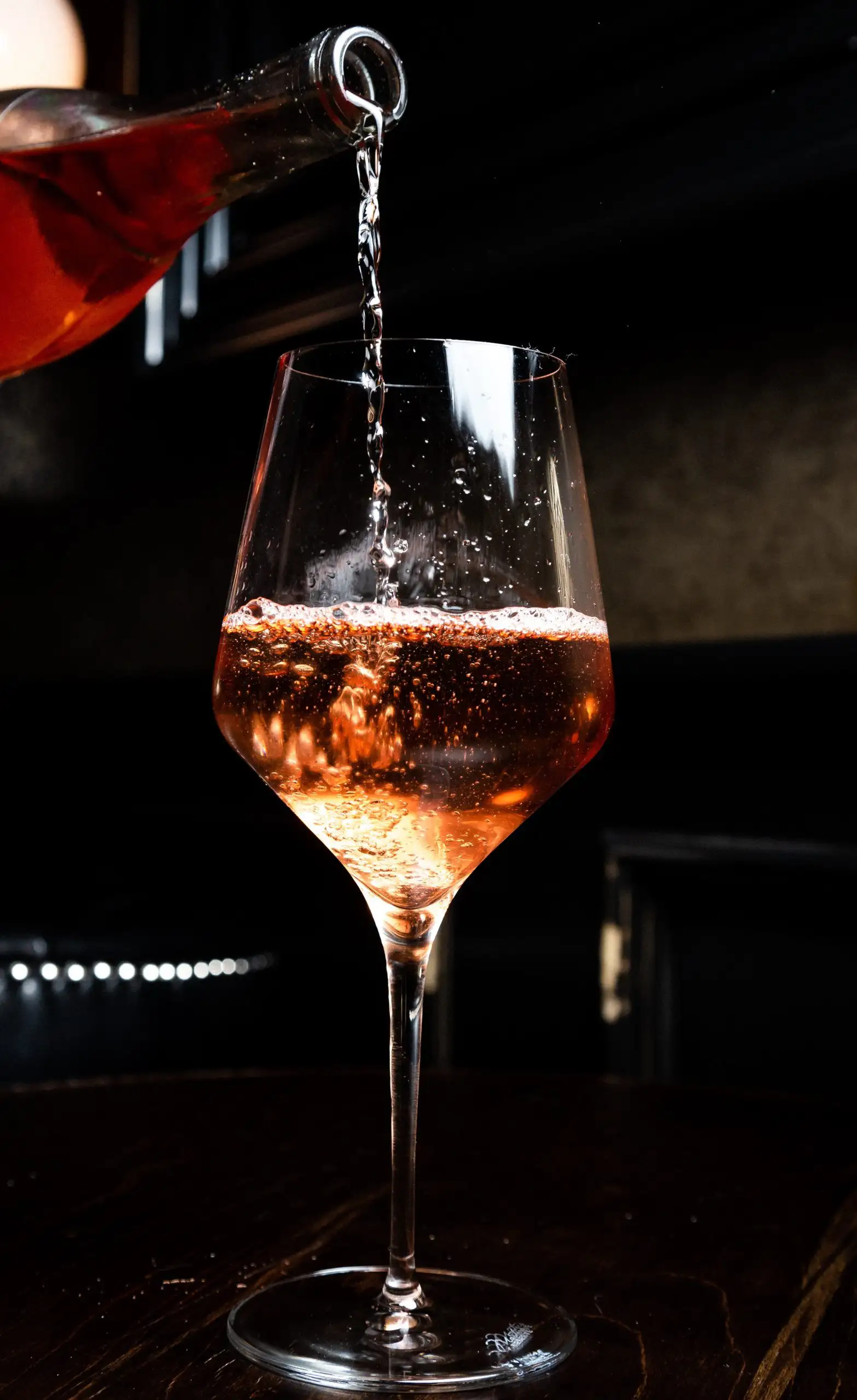 Rosé wine being poured from a bottle into a clear stemmed wine glass on a dark table.