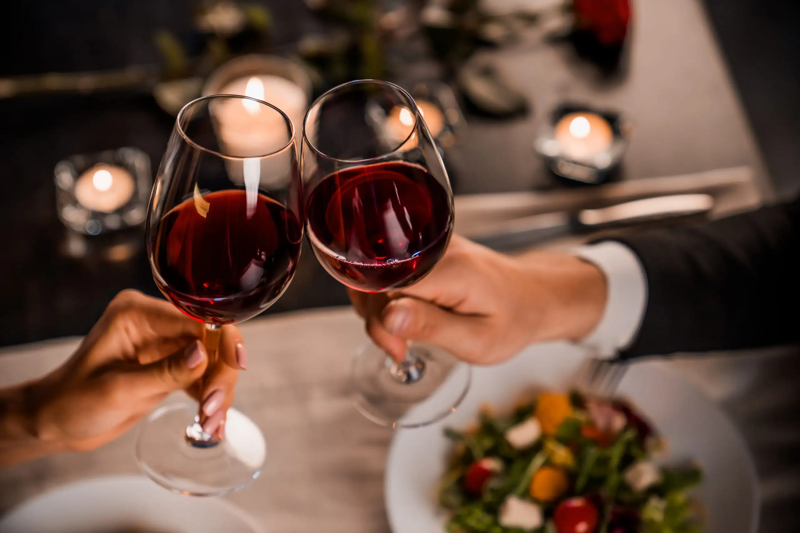 The Best Wines for Valentine's Day | Domenico Winery A bottle of red wine and a full wine glass on a table decorated with pink rose petals and red roses.