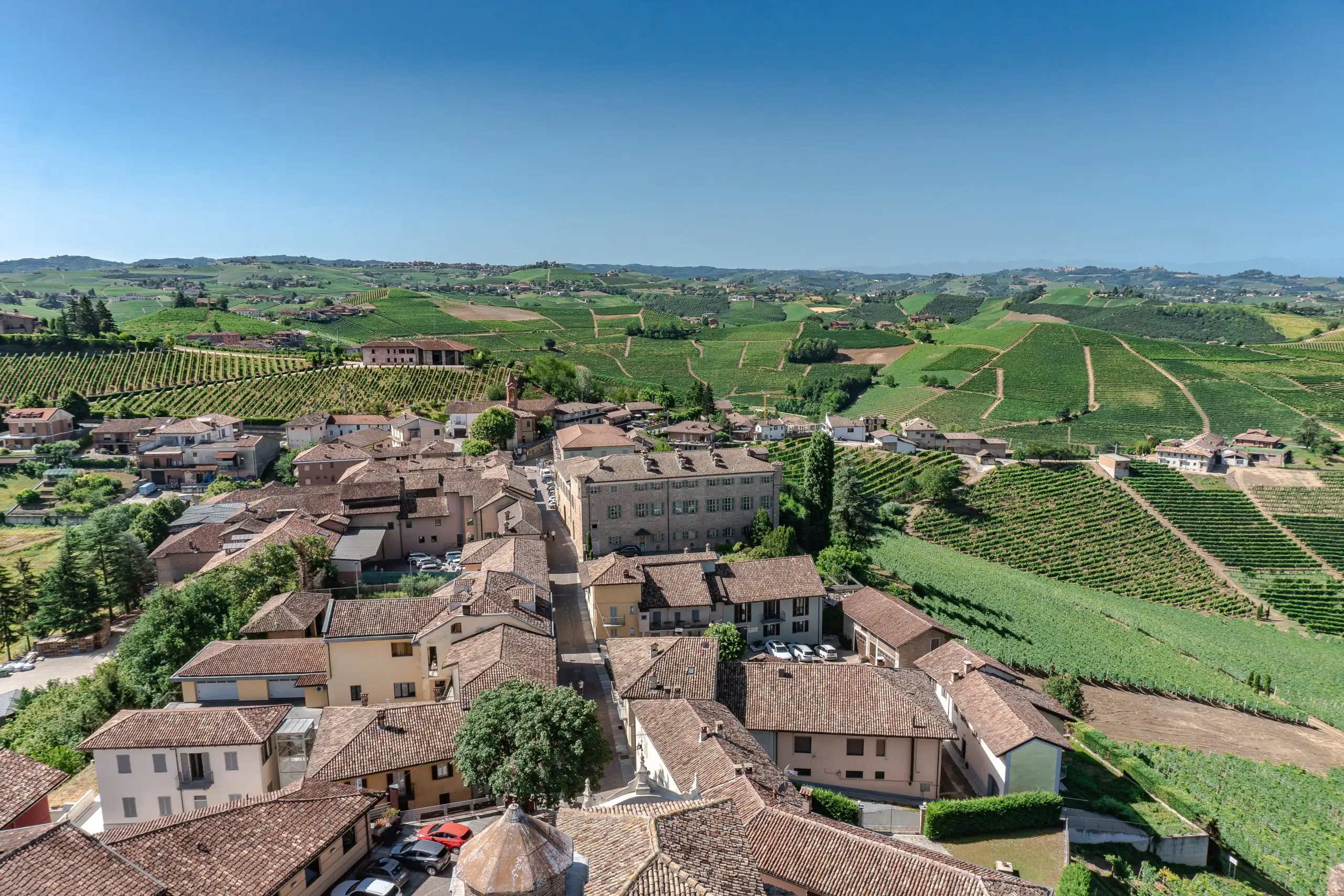 A scenic view of a village with terracotta-roofed houses, surrounded by lush vineyards and rolling hills on a sunny day.