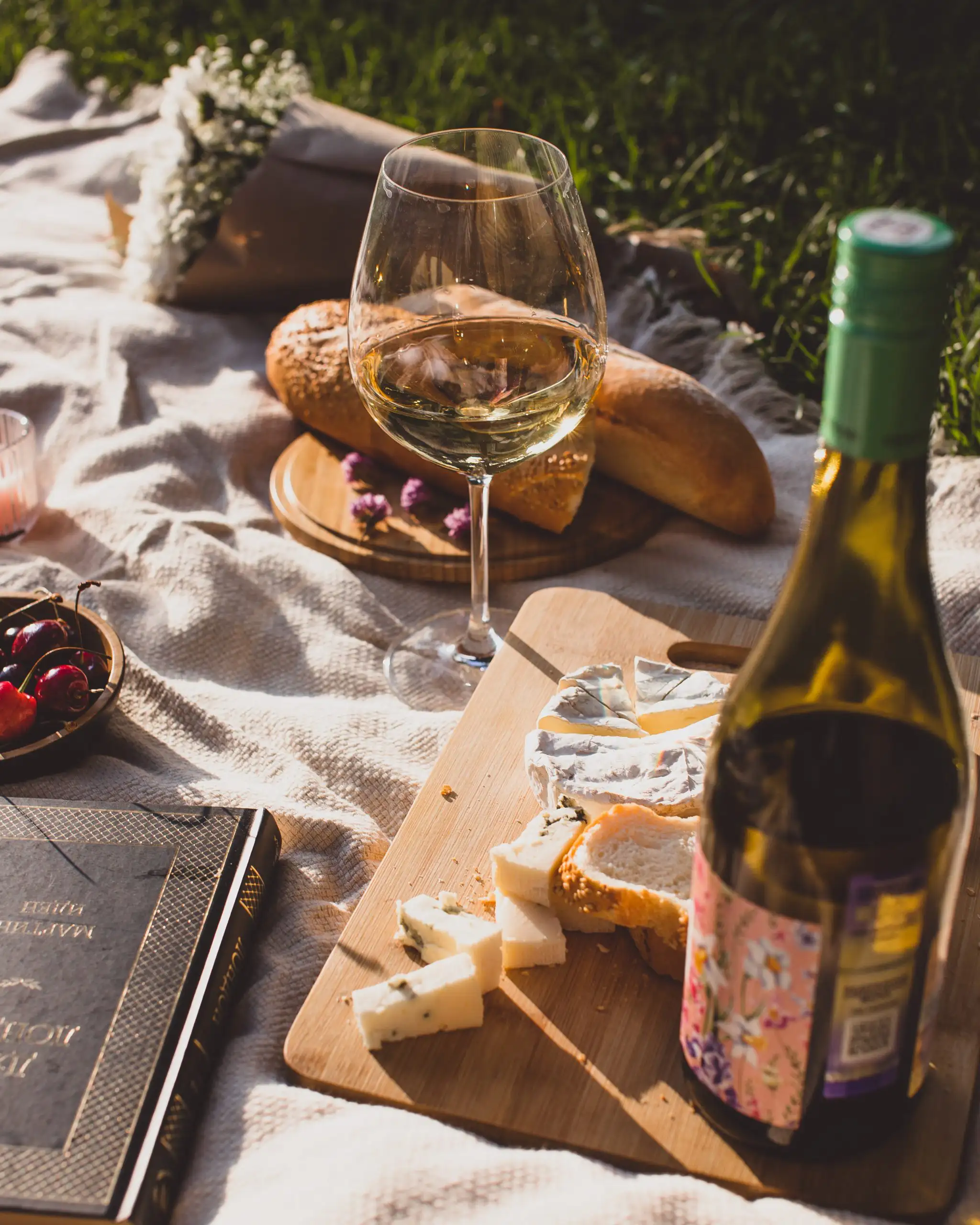 Glass of white wine on a picnic blanket with cheese, sliced bread, and a wine bottle on a wooden board.