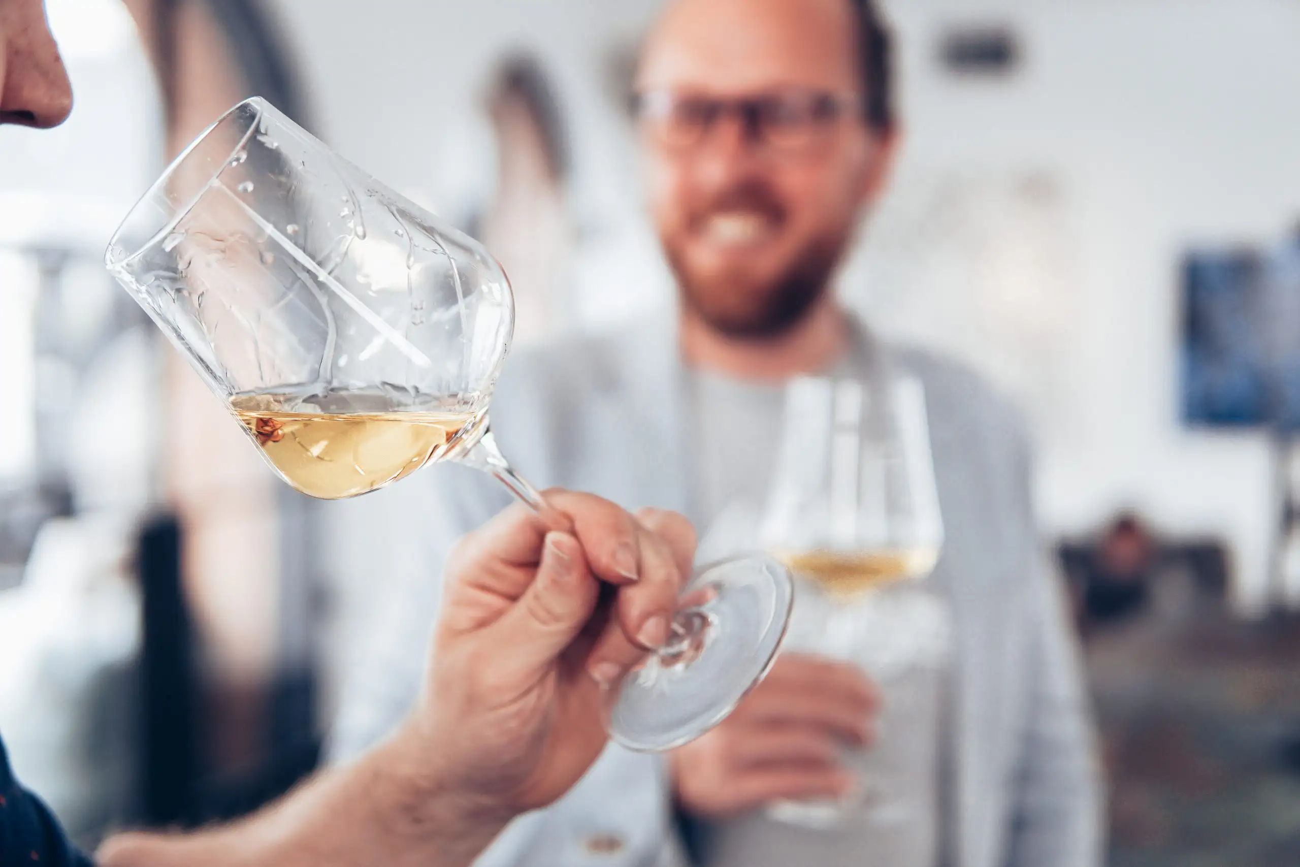 Person swirling white wine in a stemmed glass during a wine tasting while another person holds a glass in the background.