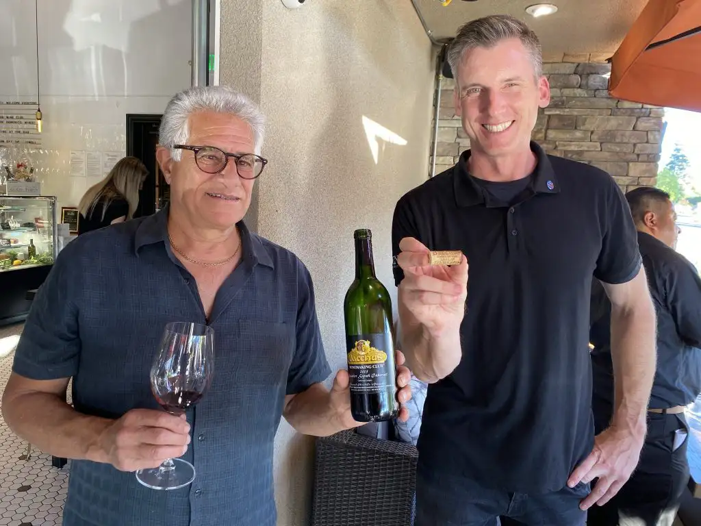 Domenico Winery Throwback Story that Will Warm your Heart! | Domenico Winery Domenick holding a bottle of red wine and a filled wine glass during a wine tasting event.