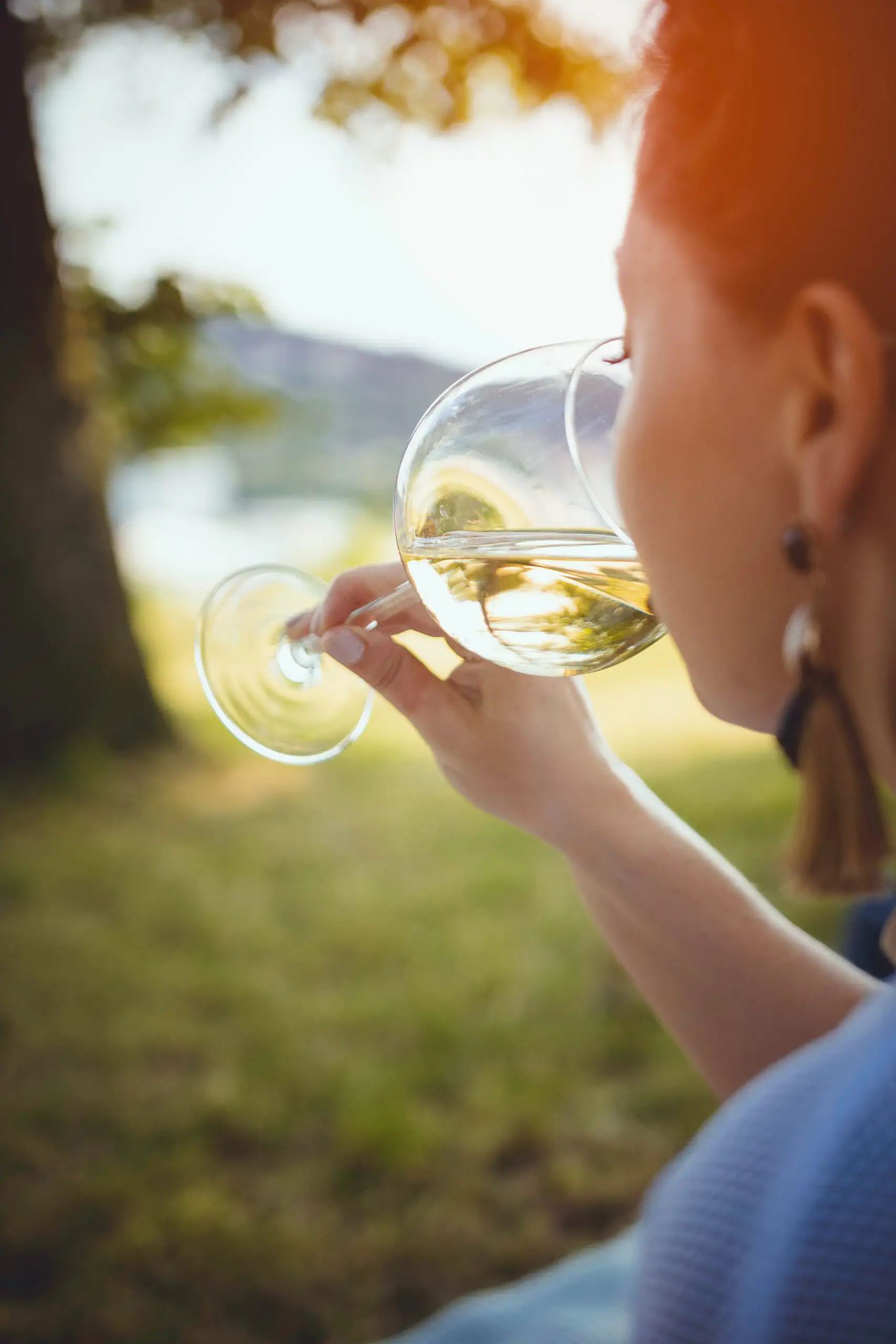 Woman holding a glass of white wine and taking a sip outdoors near a lake.